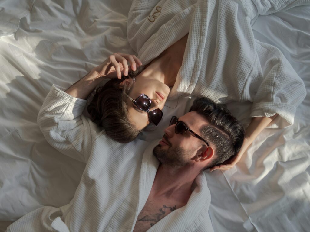 Chic couple lounging on a bed in robes and sunglasses, embracing luxury and relaxation.
