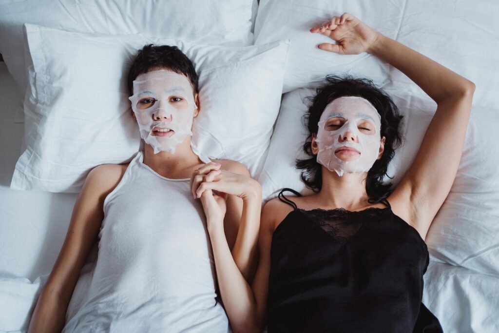 Two women relaxing with beauty masks in bed, holding hands, enjoying a self-care moment.